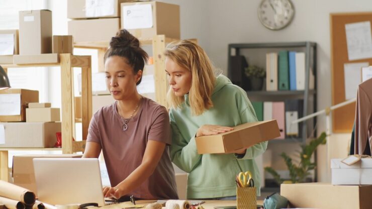Two young female entrepreneurs reviewing order packaging details in their home-based retail workspace