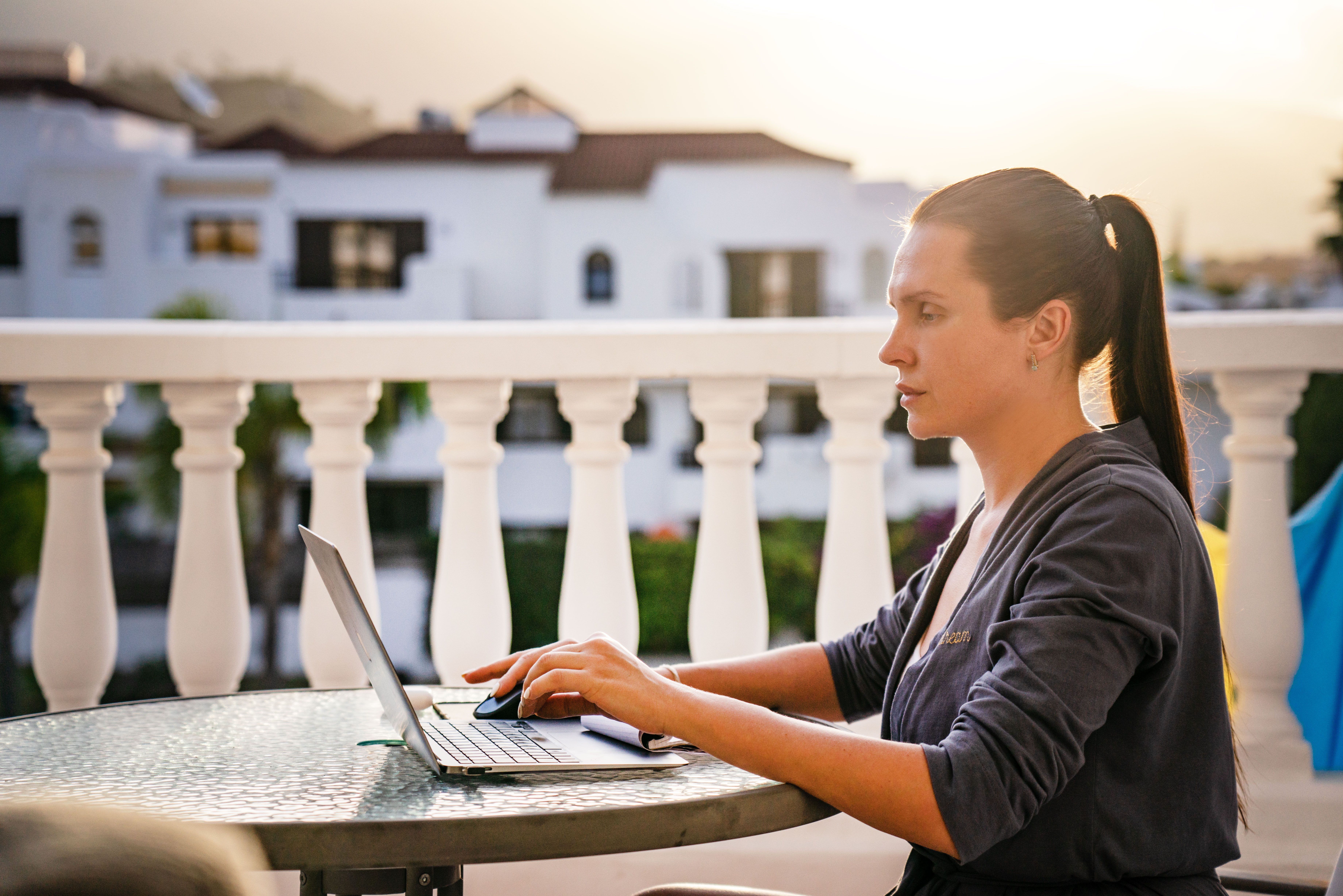 A focused woman works on her laptop outdoors on a sunny balcony